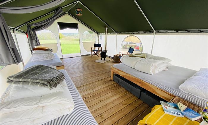 Inside a glamping tent showing a king size and two single beds, with a a log burner and chairs on a decking style floor. Background of green fields